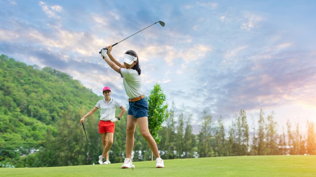 Golf ball on tee ready to be struck by woman golfer as partner watches with sunset
